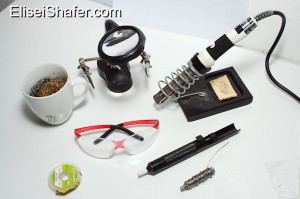 Top:Brass shavings in cup, helping hands with magnifying glass, Soldering iron with stand. Bottom:Solder wick, safety glasses, solder sucker solder.
